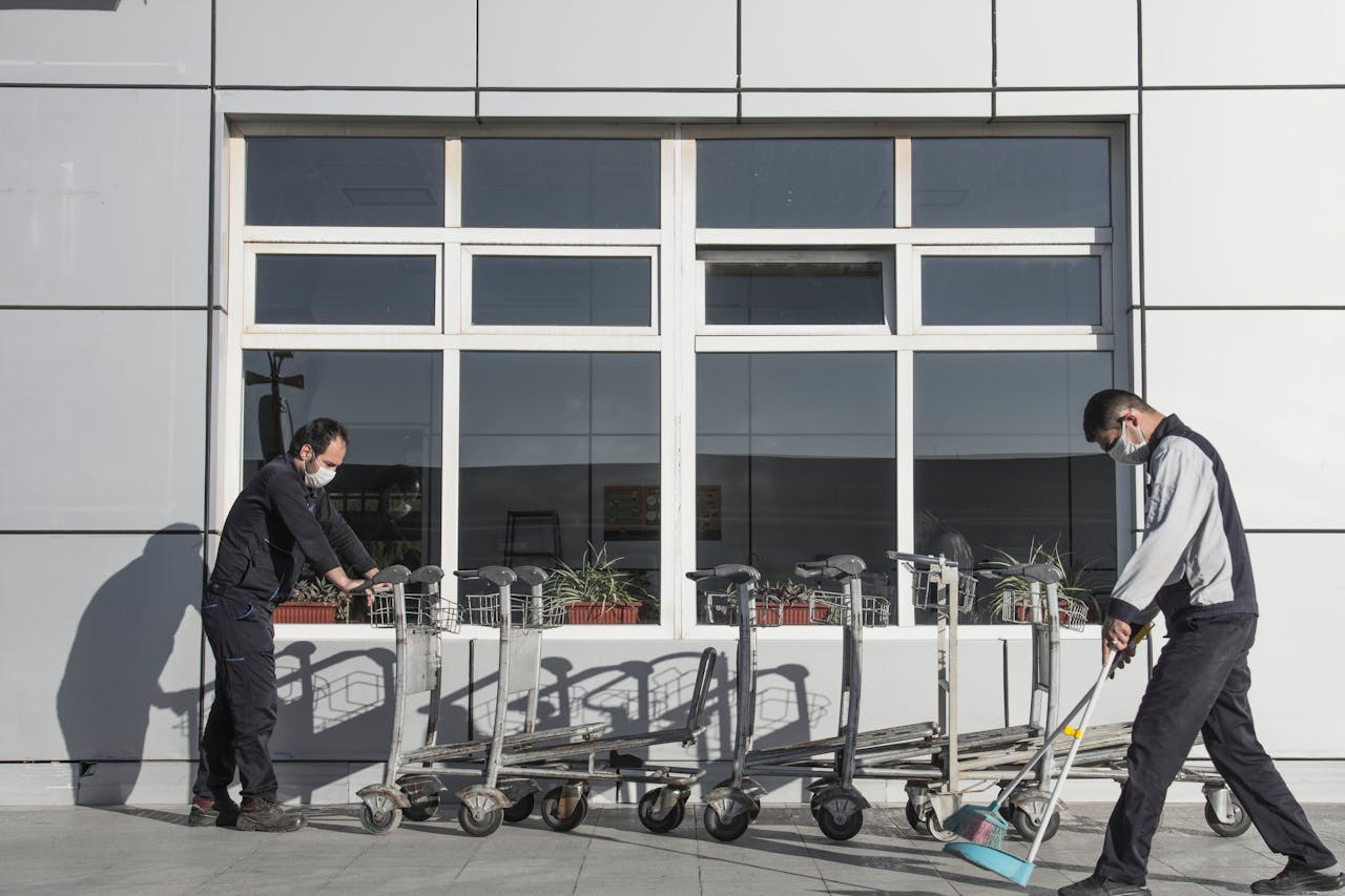 who-we-are Two masked workers clean and organize trolleys outside a modern building, highlighting industrial maintenance.